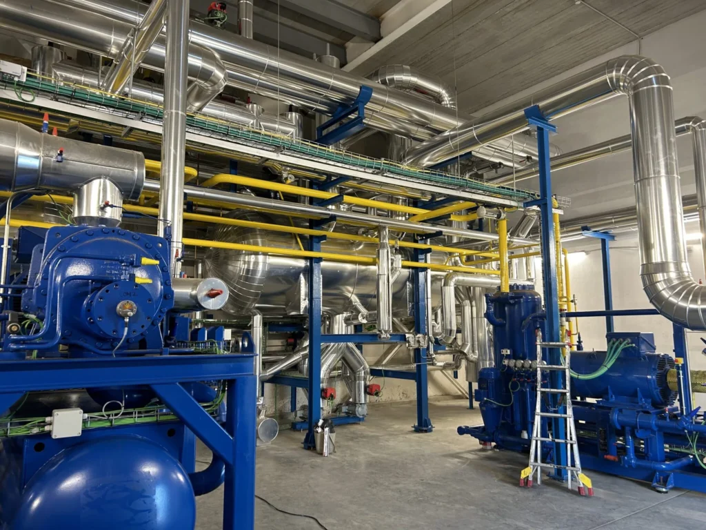 Sala de máquinas de frío industrial con compresores sobre bastidor azul y tubería aislada de gran diámetro