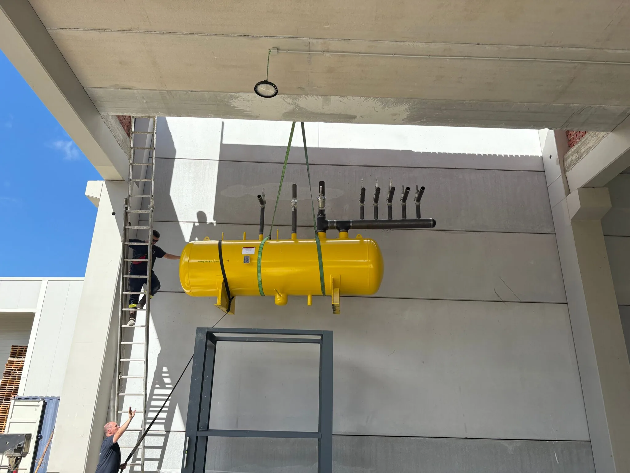 Depósito acumulador amarillo con colector de tubería, izado con eslingas durante el montaje de una planta de glicol