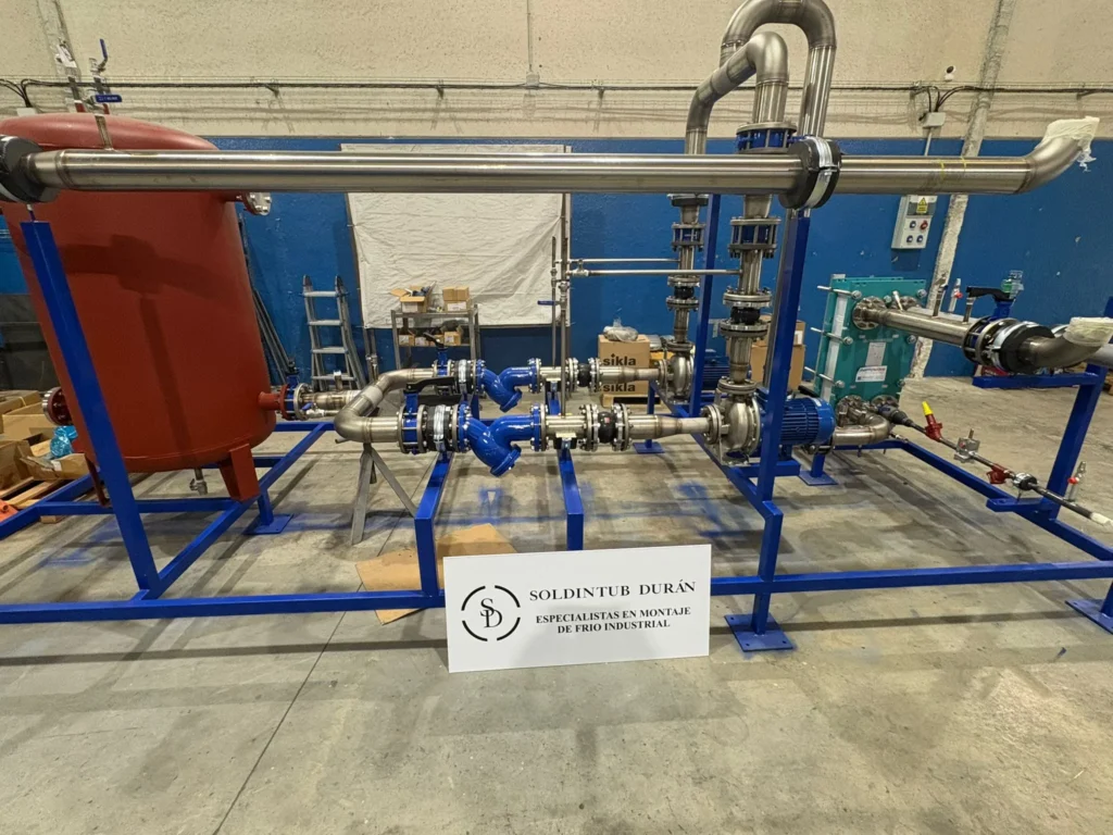 Skid prefabricado en taller con depósito rojo, bombas, intercambiador de placas y válvulas sobre bastidor azul